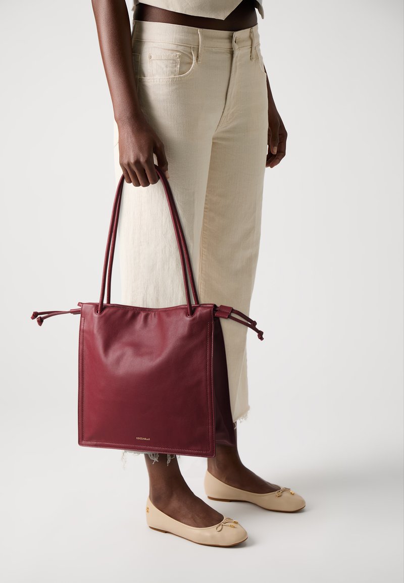 Bordeaux leren tote bag met twee lange handvatten en een trekkoordsluiting. Gecombineerd met lichtgekleurde, wijde broek en platte beige schoenen.