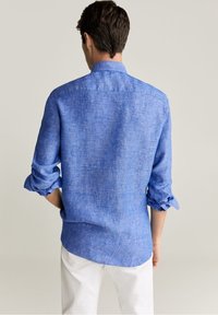 Camisa azul de lino con mangas remangadas, textura visible y sutiles variaciones de tono, combinada con pantalones blancos, vista desde atrás.