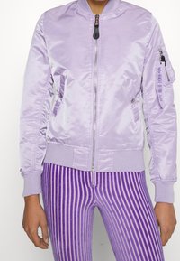 Fialová saténová bomber bunda se zipem vpředu, žebrovanými manžetami a pasem. Obsahuje boční kapsy a kapsu na rukávu se zipem s potiskem.