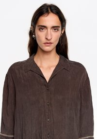 Brązowa koszula na guziki z lekkiego materiału; posiada klasyczny kołnierzyk, długie rękawy oraz teksturowane wykończenie z subtelnym przeszyciem.