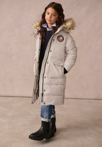 Helle graue Daunenjacke mit einer mit Fell gefütterten Kapuze, Logo-Patch, knöpffront und zwei vorderen Taschen. Kombiniert mit blauen Jeans und schwarzen Stiefeletten.