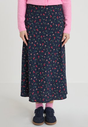 Person iført en navyblå blomsterprintet midinederdel kombineret med lyserøde sokker, navyblå sneakers og en lyserød sweater, stående indendørs på et lyst gulv.
