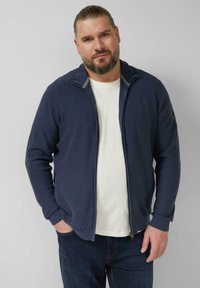 Marineblauw jack met rits, geribbelde kraag en manchetten, met een gestructureerd ontwerp en een logo op de linker schouder, gedragen over een wit T-shirt.