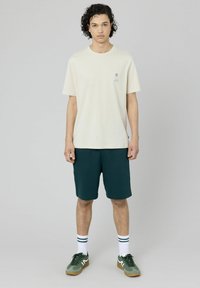 T-shirt beige à manches courtes avec un petit graphisme, associé à un short vert foncé. Porté avec des chaussettes blanches à rayures vertes et des chaussures vertes.