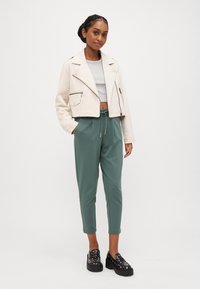 Kuttet beige jakke med glidelås, grå crop top, grønne bukser med snøring og svarte platå sko med sølv aksenter.