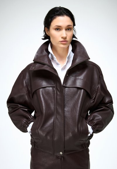 Rövid sötét hajú nő sötétbarna bőrdzsekit visel fehér galléros ing fölött, kezei a zsebében, semleges arckifejezés, egyszínű világos háttér.