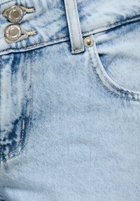 Gros plan sur un jean en denim bleu clair montrant la poche avant, la passants de ceinture, la fermeture à double bouton en métal et les coutures détaillées.