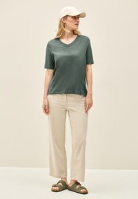 Mørkegrøn kortærmet V-hals T-shirt, beige afslappede bukser, og mørkegrønne sandaler med to remme, båret med en lysfarvet kasket.