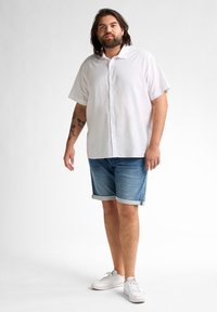Lichte, kortebloezige witte overhemd met knopen, gecombineerd met opgerolde denim shorts. Draagt witte sneakers, voor een neutrale achtergrond.