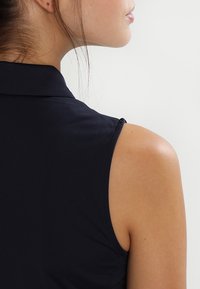 Marinblå ärmlös polo-top som visar en strukturerad krage, slät textur och rena sömnadsdetaljer, sedd bakifrån.