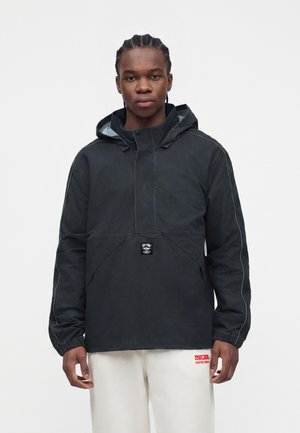 Jeune homme aux cheveux tressés portant une veste à capuche noire et un pantalon blanc avec un texte rouge sur un fond clair uni.