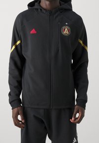 Svart zip-up hoodie i slät tyg, med ett gult streck och rosa Adidas-logotyp, samt Atlanta United FC-emblem på bröstet.