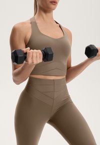 Brązowy, dopasowany sportowy top w stylu crop oraz pasujące legginsy z wysokim stanem. Materiał wydaje się gładki z teksturowanymi liniowymi wzorami. Czarne sześciokątne hantle trzymane w dłoniach.