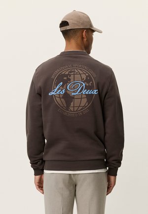Sudadera marrón oscuro con un gráfico redondo que muestra un globo terráqueo y "Les Deux" en azul claro, con puños y dobladillo acanalados. Combinada con pantalones gris claro.