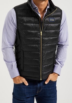 Mann trägt eine schwarze gesteppte Weste mit Reißverschluss über einem fliederfarbenen Hemd mit Knöpfen und dunkelblauen Jeans, Hände in den Taschen. Polo Club Logo auf der Brust der Weste.