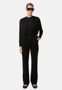 Schwarzer Cardigan mit Knöpfen, eng anliegendes schwarzes Oberteil und weite schwarze Hosen, kombiniert mit flachen schwarzen Schuhen und übergroßen schwarzen Sonnenbrillen.