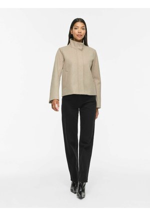 Kvinde iført en beige lynlåstrøje, sorte lige bukser og sorte ankelstøvler, stående mod en ensfarvet hvid baggrund.