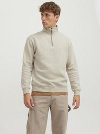 Hellbeige Zip-up-Sweatshirt mit hohem Kragen, weichem Textil und gerippten Bündchen, über einem braunen Shirt getragen. Das Modell trägt hellbeige Hosen.