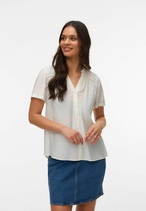 Femme souriante aux longs cheveux noirs portant un chemisier blanc à manches courtes avec des détails en dentelle et une jupe en jean bleue, debout devant un fond blanc.