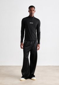 Mann iført svart Calvin Klein langermet topp, vide svarte bukser med glidelåslommer, og sko med hvite såler som står på beige gulv.