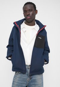 Navyblå jakke med sort brystlomme og røde detaljer, lavet af let materiale. Har hætte og løs pasform, bæres over en hvid skjorte.