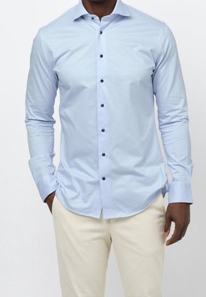 Mies, joka pukeutunut vaaleansiniseen nappipaitaan mustilla napeilla ja beigenvärisiin housuihin, seisoo yhdellä kädellä taskussa.