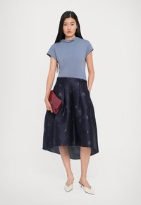 Woman stands with a pale blue top, navy patterned midi skirt, white heels, and holds a red clutch against a plain white background.