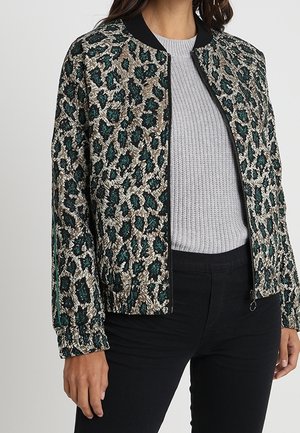 Femme portant une veste bomber texturée à motif léopard avec des taches vertes et beiges, par-dessus un pull en maille gris clair et un pantalon noir.