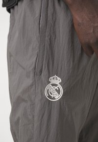 Pantalones ligeros gris claro con una textura suave, que presentan un detallado logotipo bordado en blanco con una corona en la parte inferior de la pierna izquierda.