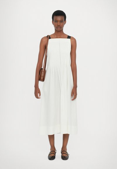Weißes ärmelloses Kleid mit Falten, quadratischem Ausschnitt und schwarzen Trägern. Kombiniert mit schwarzen Ballerinas und einer braunen Umhängetasche. Einfaches Design.