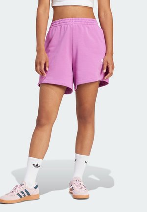 Person iført lyserøde træningsshorts, hvide crew-sokker med sort logo og lyserøde sneakers, stående mod en lys baggrund.