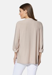 Beige Langarmtop mit umgeschlagenen Bündchen, glatter Textur, lockerer Passform und seitlichen Schlitzen, von hinten vor einem schlichten Hintergrund betrachtet.