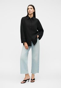 Femme debout portant une chemise noire ample boutonnée, un jean large bleu clair avec des ourlets effilochés et des sandales noires à talons.