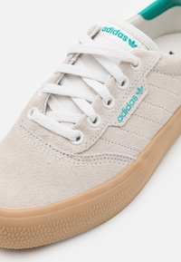Närbild av beige mockaskor från Adidas med vita snören, teal-färgad logotyp på tungan och sidan samt gummisula.