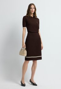 Vrouw staand in een bruin kortärmlig topje en geplooide rok met witte bies, ze houdt een beige handtas vast en draagt zwarte hakken tegen een witte achtergrond.