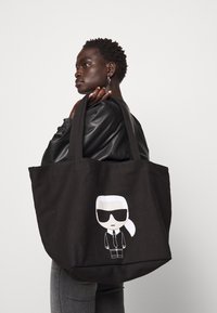 Osoba v černé kožené bundičce nese velkou černou tašku s kreslenou postavičkou v slunečních brýlích a s bílými vlasy svázanými dozadu.