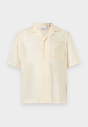 Homme portant une chemise jaune pâle à manches courtes boutonnée, un short beige avec une ceinture noire, et des sandales noires, debout devant un fond blanc.