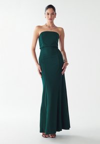 Nais, kes kannab õlavöödeta maani tumeroheline kleit koos kuldsete ehtetega ja mustade avatud varvastega kontsakingadega, seisab ühtlasel valgel taustal.