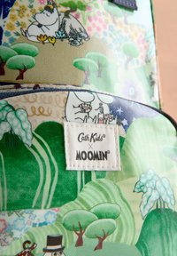 Barvna tkanina z liki Moomin, drevesi in hribi. Vključuje belo oznako z napisom "Cath Kids x MOOMIN" v črni barvi.