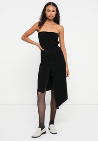 Top negru fără bretele, din material canelat, cu un detaliu cu fundă, asortat cu o fustă asimetrică neagră, cu o deschidere pe o parte. Modelul poartă ciorapi negri transparenți și pantofi deschiși.