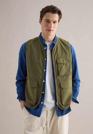 Jonge man die een groen werktuigvest draagt over een blauw spijkershirt en wit T-shirt, staand met handen in de zakken tegen een effen achtergrond.