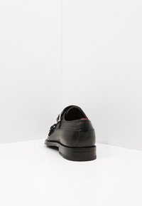 Chaussure en cuir noir avec une finition texturée, maintenue par une boucle, et une semelle plate robuste. La vue arrière présente des accents de design minimalistes.