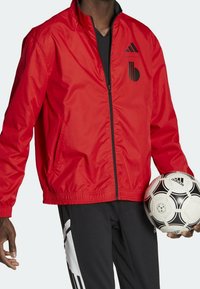 Veste légère rouge avec col montant, logo noir, deux poches latérales et ourlet élastiqué, accompagnée d'un ballon de football noir.