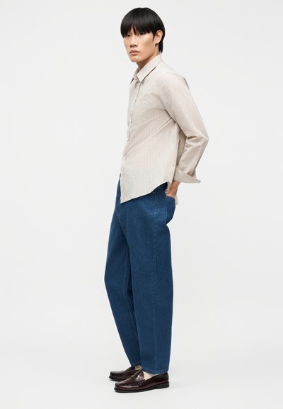 Νεαρό άτομο όρθιο πλάγια, φορώντας μπεζ μακρυμάνικο πουκάμισο, φαρδιά μπλε τζιν, λευκές κάλτσες και καφέ-σκούρα μοκασίνια, με το ένα χέρι στην τσέπη.