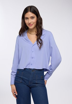 Cropped blouse di un leggero viola con frontale a bottoni, colletto e maniche lunghe. Indossato con jeans scuri a vita alta dotati di tasche utilitarie.