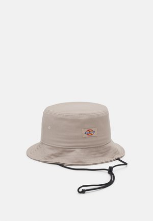 Beige Bucket Hat aus strapazierfähigem Stoff mit einer runden Krone, breiter Krempe und verstellbarem schwarzen Kinnband. Verfügt über ein Logo-Patch an der Seite.