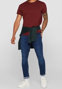 Hombre con una camiseta burdeos, jeans oscuros con los puños arremangados, zapatillas blancas y una sudadera oscura atada a la cintura.