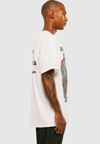 Hvid bomulds t-shirt med korte ærmer, med et farverigt grafisk print på siden, der viser en person, og fed tekst på ryggen.