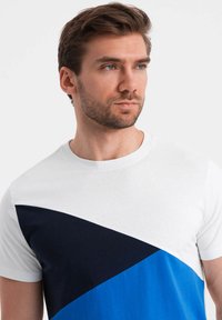 Tričko s krátkým rukávem v bílé barvě, s diagonálním designem v námořnické modré a modré části. Vyrobeno z bavlny, má kulatý výstřih a hladkou texturu.