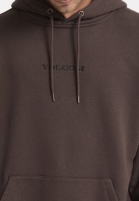 Sweatshirt à capuche marron en matériau doux, avec un logo "VOLCOM" brodé en noir, des cordons de serrage à embouts métalliques et une poche frontale.